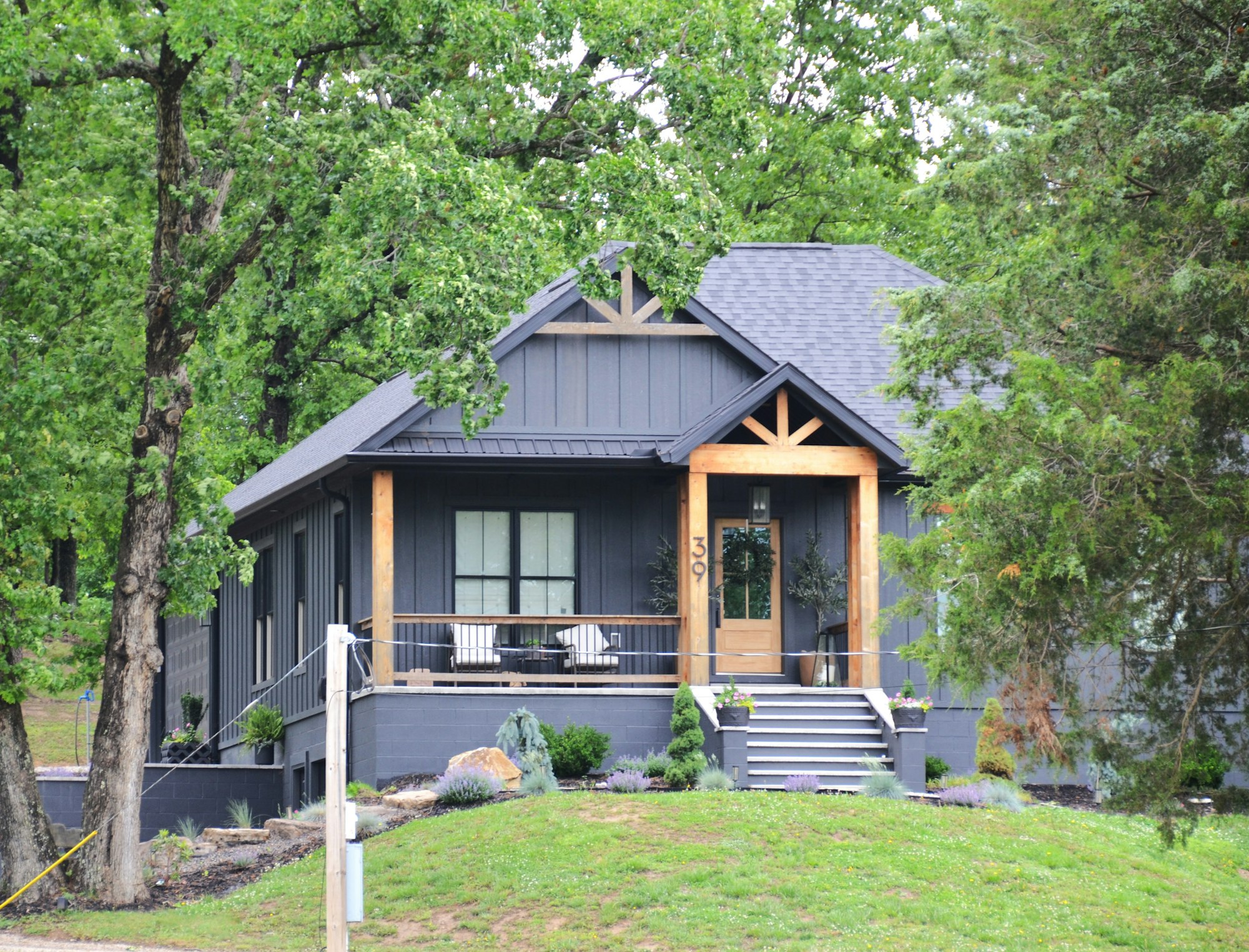 A dark gray home with a wooden front porch among green trees
