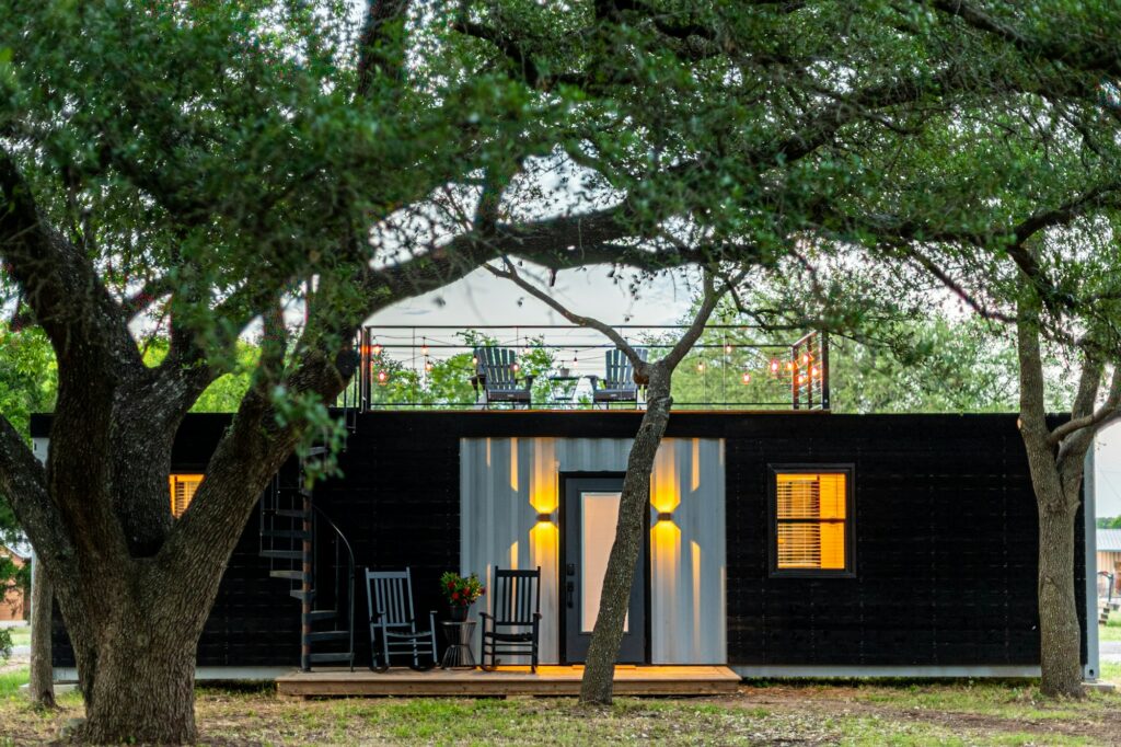 A small black home with a porch under large oak trees
